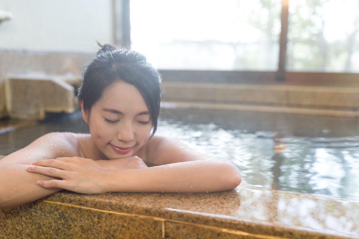 A woman rests beside a steaming indoor Tokyo onsen pool, eyes closed, surrounded by tiled walls and warm mineral water.