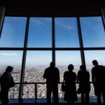 A business executive reviews documents at a Tokyo headquarters office, with a city skyline visible through the window.