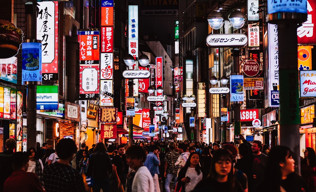 A crowded neon-lit street in Japan at night, with colorful signs and storefronts lining the sidewalk as shoppers pass by.
