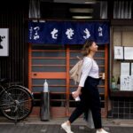 A woman dressed in loose natural Japanese fabrics walks through a shaded Tokyo street on a hot August afternoon